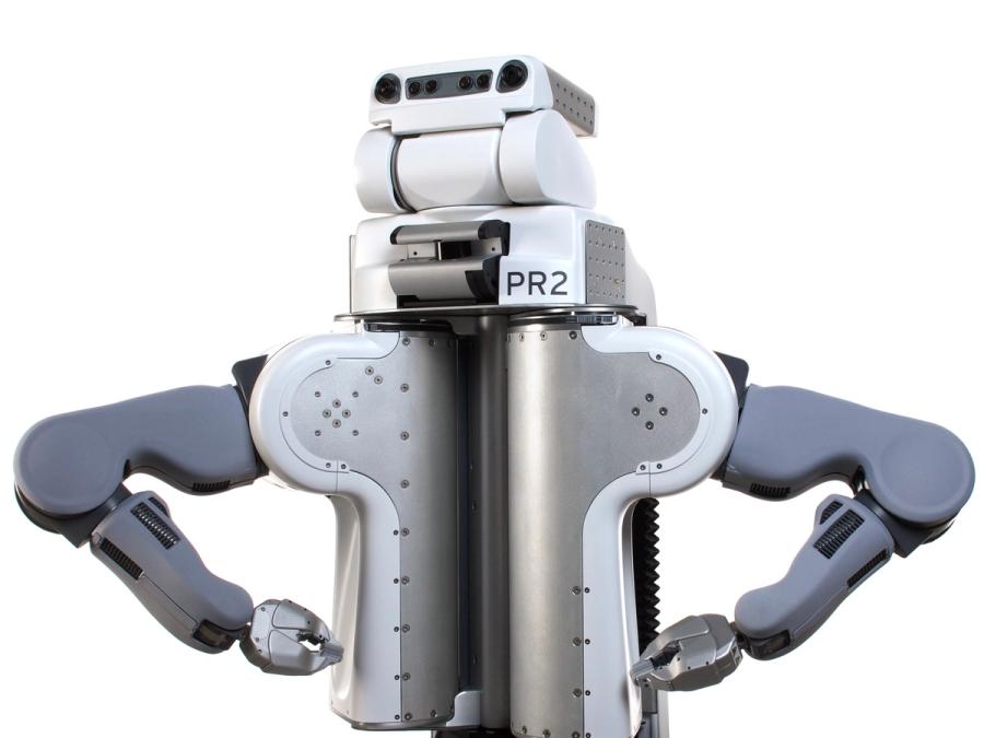 PR2's white plastic and metal torso holds three stacked sections, the bottom which says PR2 and the top of which has two eye cameras and a black bar with sensors. The robots arms, with gripper end effectors, are posed on it's hips.