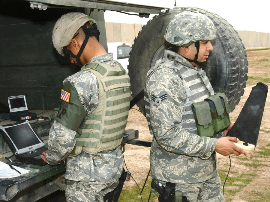 Two American soldiers in military gear stand at the back of a tank. One looks at a computer, and the other holds a piece of equipment.