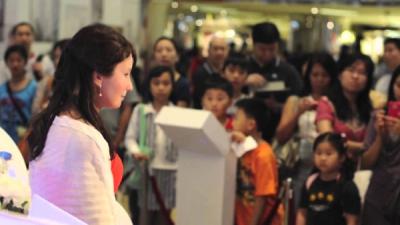 Female lifelike android wears a white robe and sits at a department store while a crowd of adults and children stare at it.