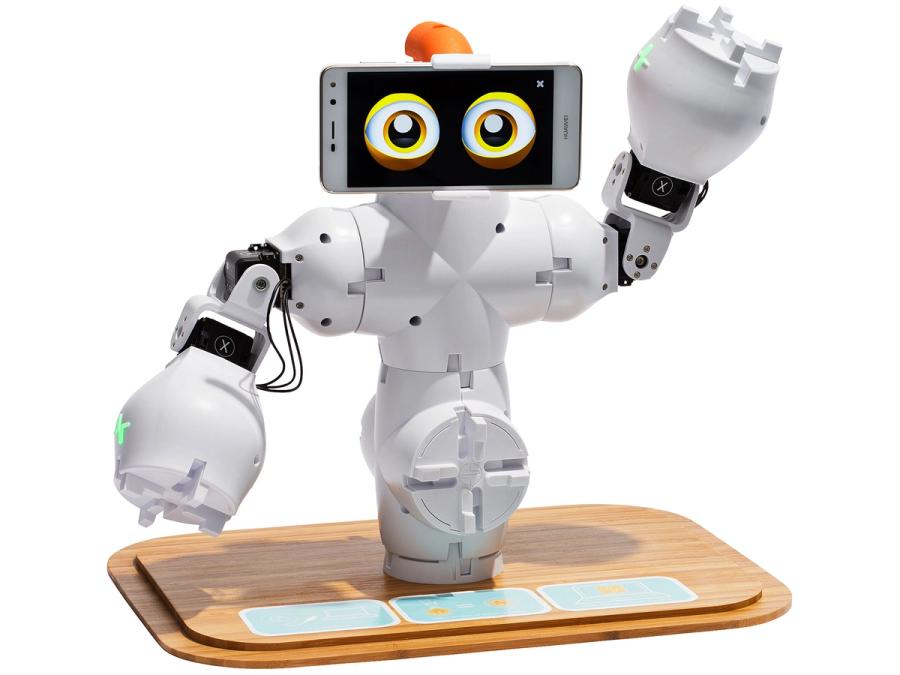 A modular white robot with a small torso, arms and a phone displaying expressive eyes sits on a wooden tabletop base.