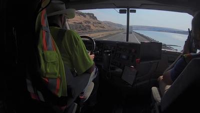 A man wearing a yellow shirt and a hat sits on the driver seat of an autonomous truck driving itself on a road.