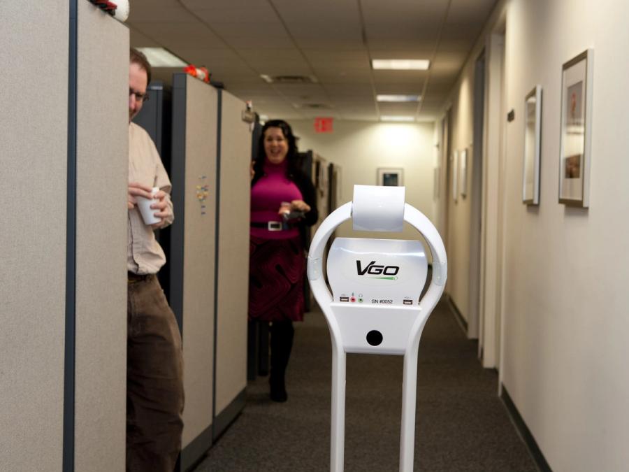 Two people smile at the telepresence robot which moves through the hallway of an office.