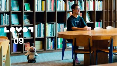 LOVOT roaming at a modern office where a man sits at a wooden desk behind bookshelves.