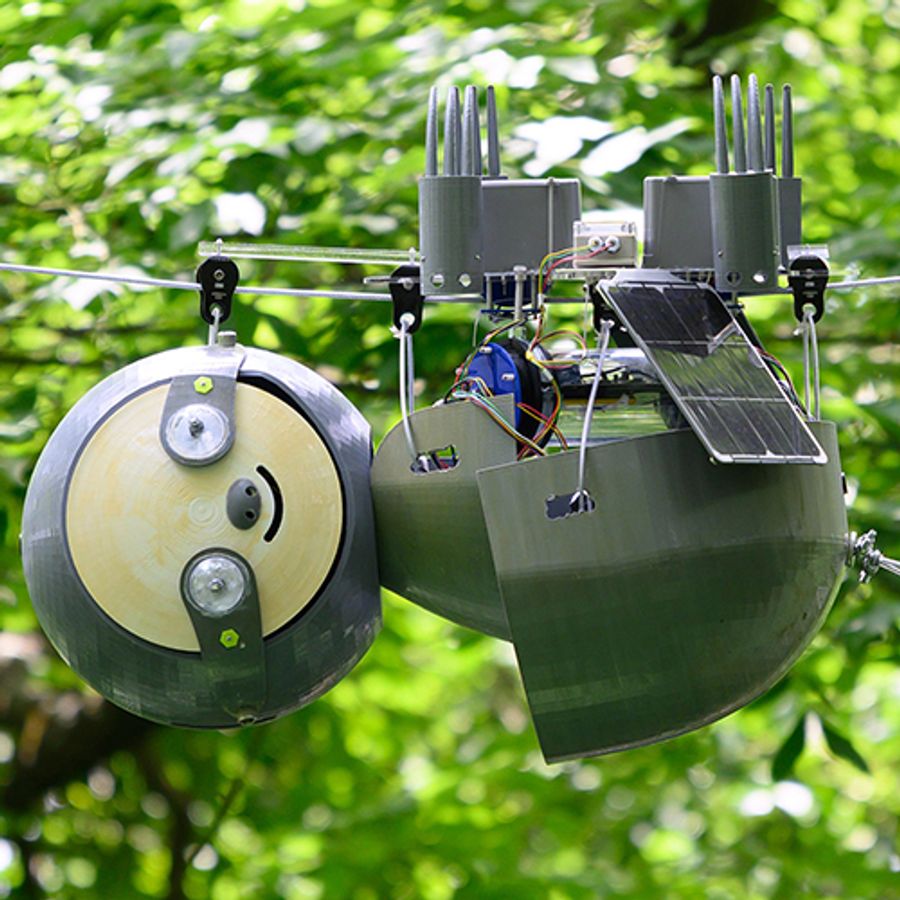 A robotic sloth hangs upside down from a wire amid green leafy trees outside. It has a solar panel and other electronics.