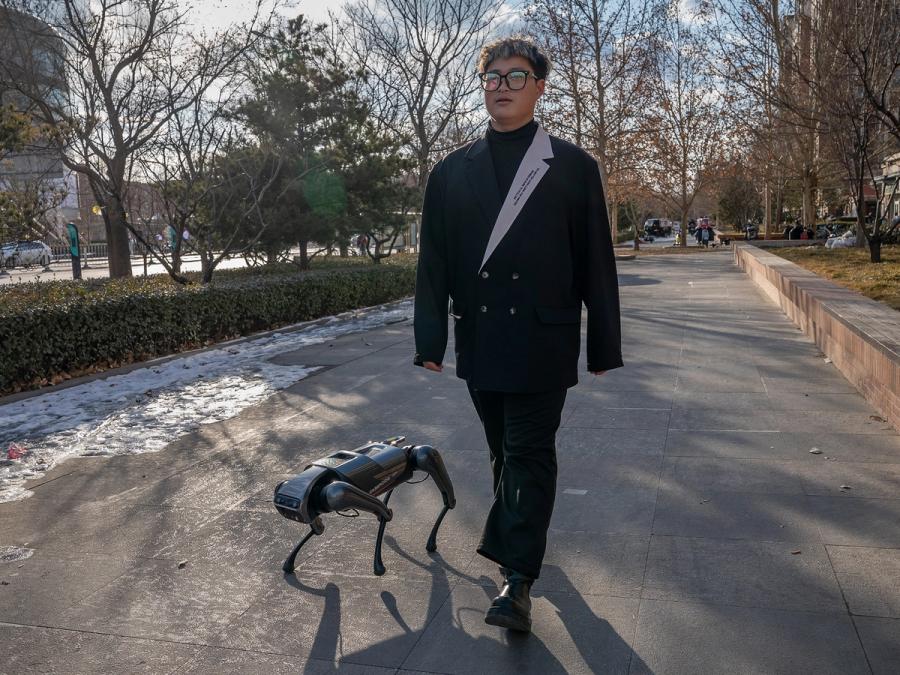 A man in glases and a fancy suit walks down a street with a small CyberDog quadruped robot by his side.