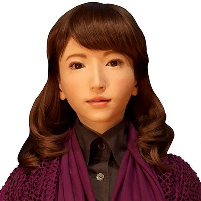 Erica, a highly realistic humanoid female robot with golden skin, almond shaped eyes, and shoulder length brown hair wears a black button up shirt and purple sweater.