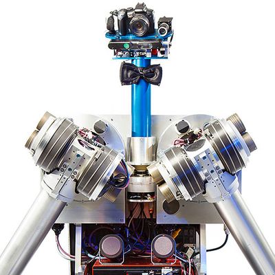 Close-up of the robot's torso, two articulated arms, blue pipe-like neck with a black bowtie on it, and a platform for a head that includes a camera system.