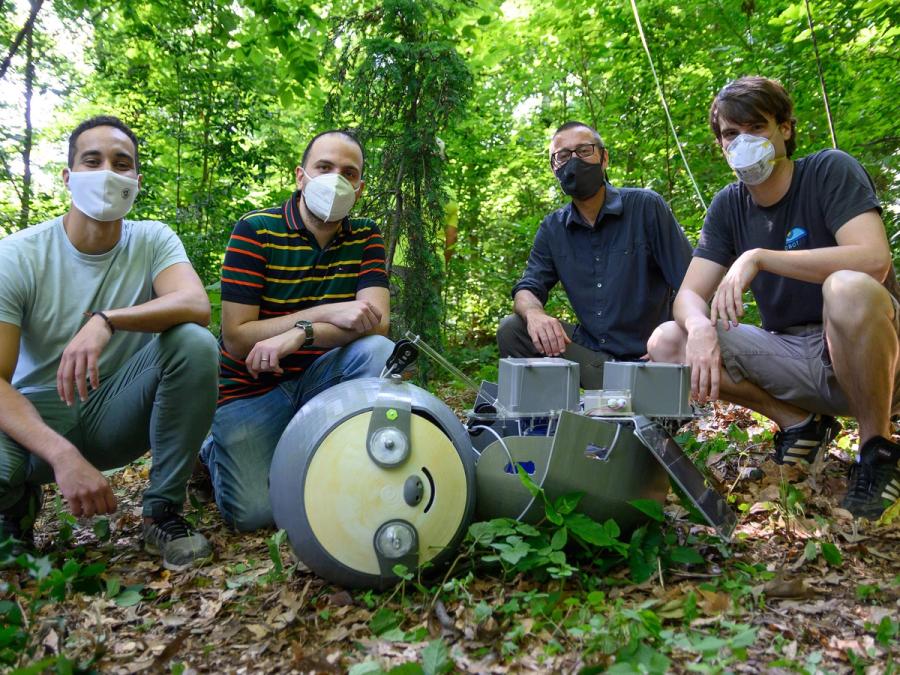 Four people in masks squat outside behind a large sloth robot.