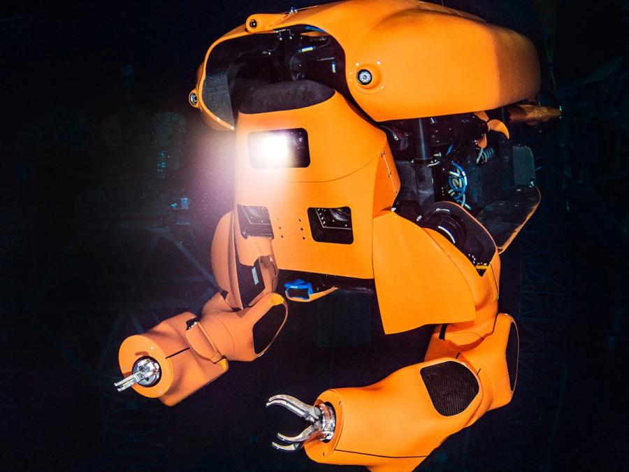 An orange half humanoid submarine robot with gripper hands shines a light underwater.