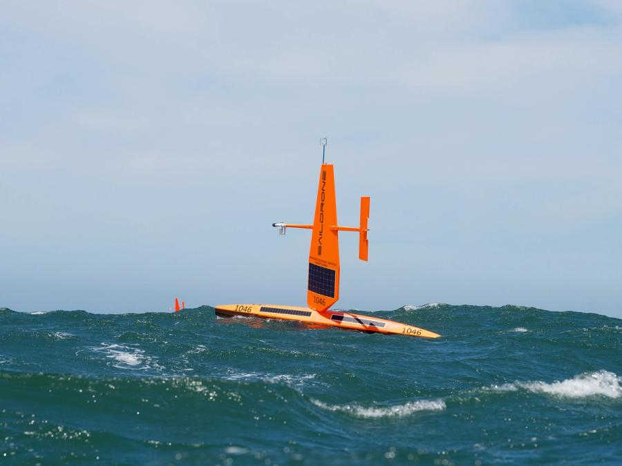 An orange uncrewed surface vehicle consisting of a horizontal unit on the water, with a large sail with solar panels rising vertically, and a rudder running parallel to the sea.