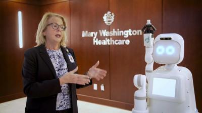 A white humanoid robot with a gripper on an articulated hand stands next to a blonde woman in glasses, in front of a sign that says Mary Washington Healthcare.