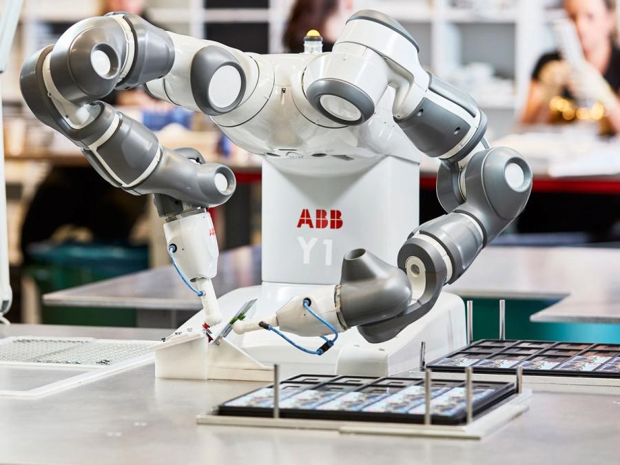 A tabletop industrial robot with a white base and torso sports two grey industrial arms. It's end effectors are a single finger each, which hold a small square item, from a tray of items on the table in front of it.