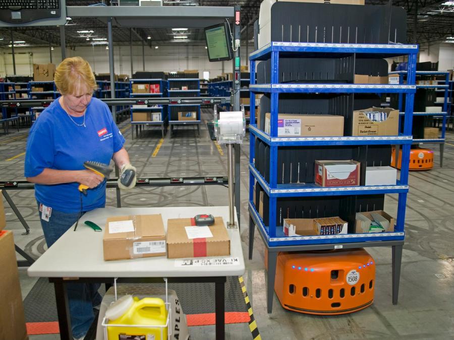 A woman stands at a table scanning items that have been brought by an orange robot carrying shelves of items.