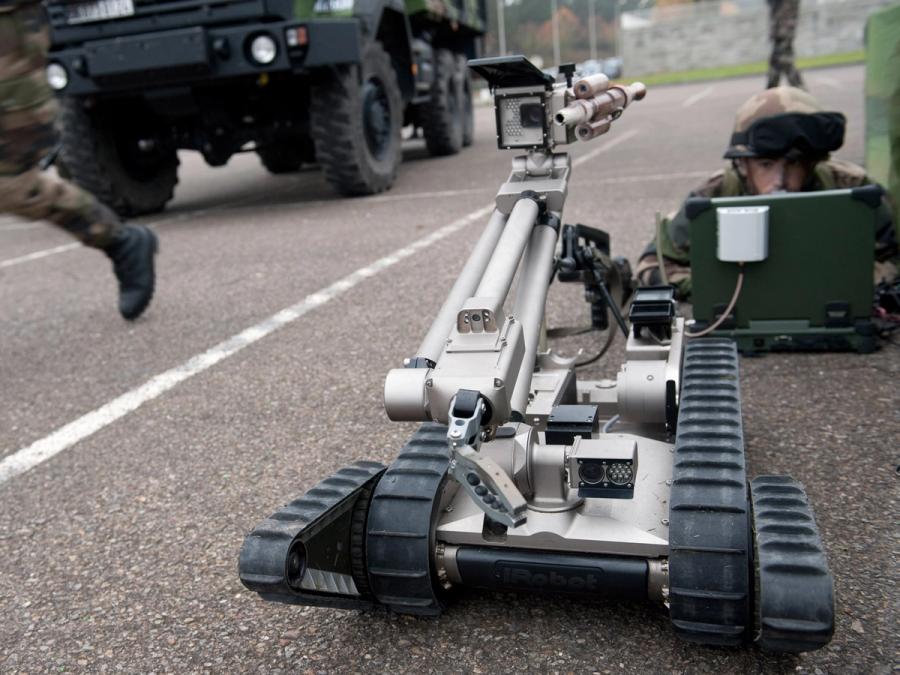 A man in military garb lies on a street looking at a computer. The PackBot is in front of him, and there is a tank to the side.