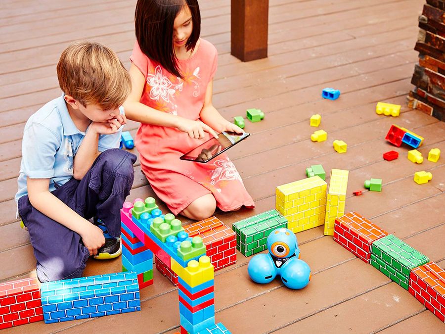 Two kids, a boy in a blue shirt and a girl in a red dress, sit on the floor and use an iPad to operate a little orange and blue mobile robot called Dash through a maze of cardboard bricks.