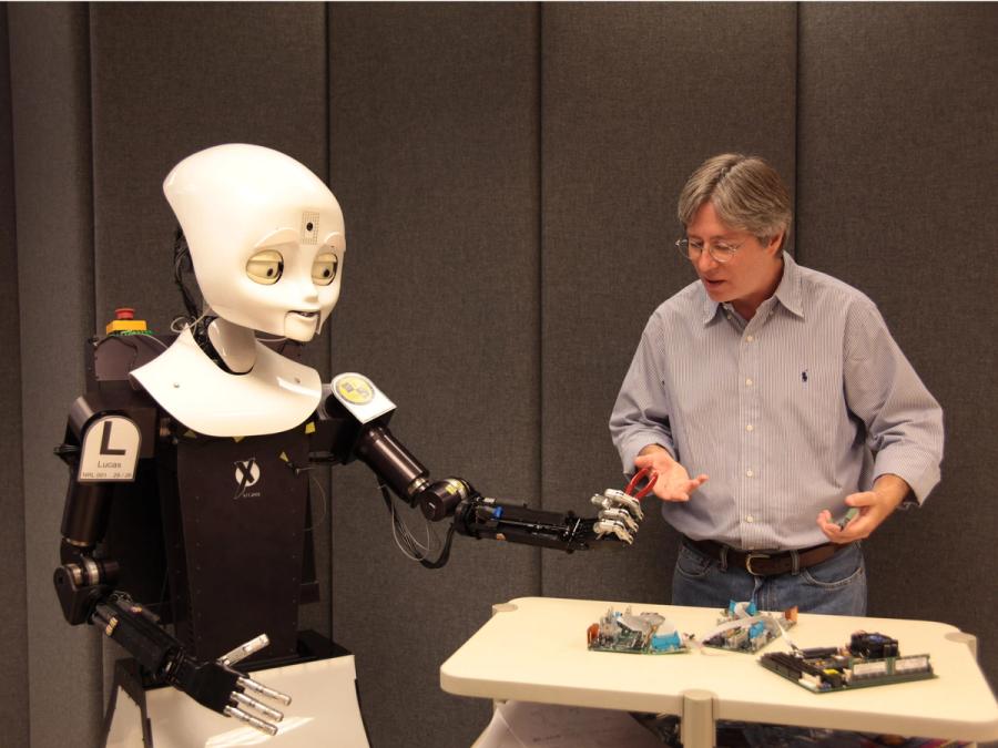 Octavia stands at a table holding an object that a man is also gripping. The robot is the same height as the human.