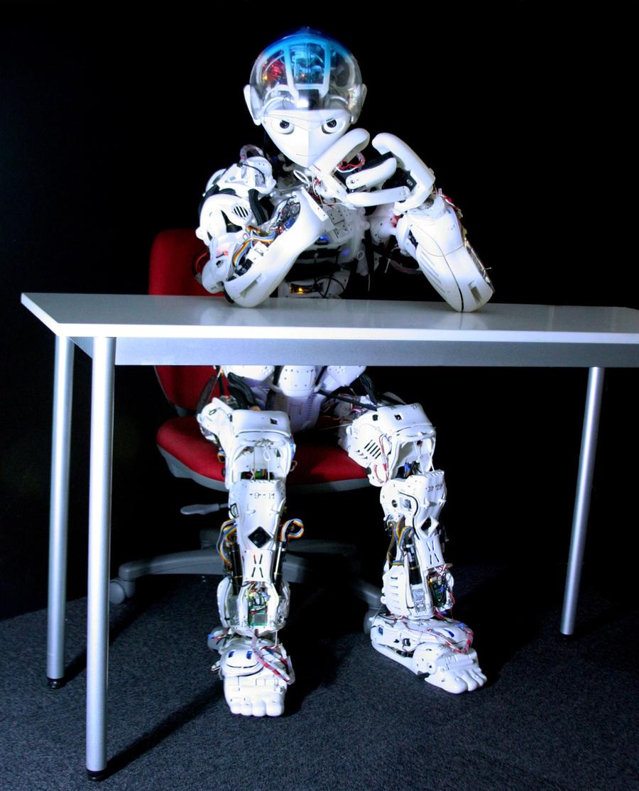 The robot is seated and posed at a table with it's hands clasped as it looks at the camera.