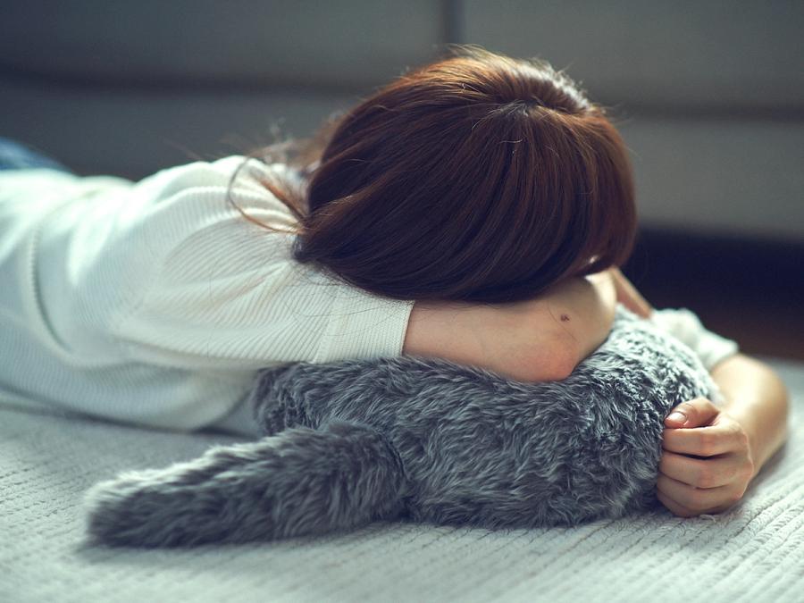 A dark haired woman in a white shirt rests her head on a round cushion with a tail, covered in soft looking gray fur.