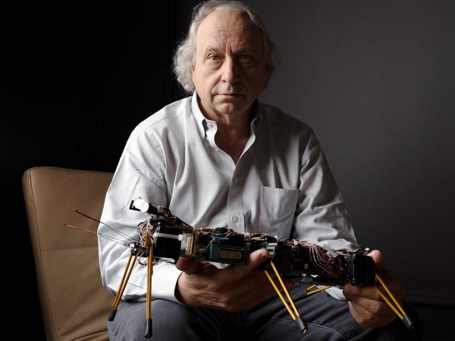 A serious man with gray hair sits holding a large robot which has a rectangular body and six simple legs.