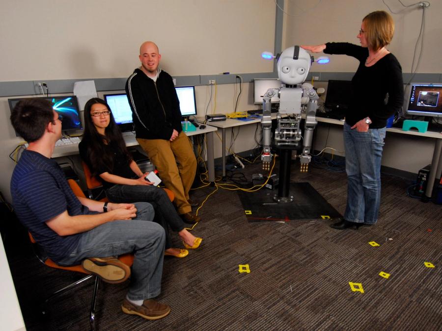 Four people hang out with Simon the robot in a computer lab.