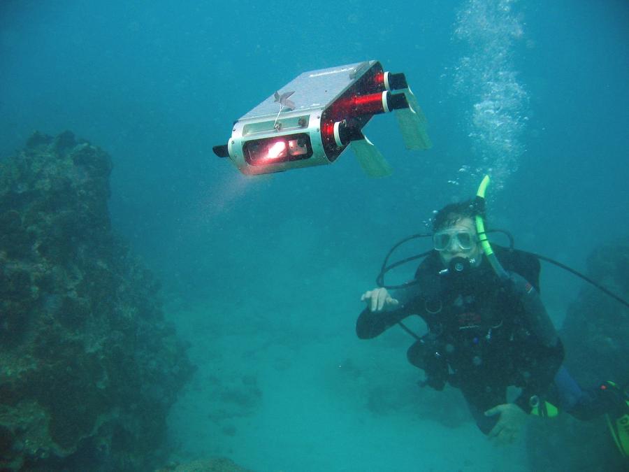 A rectangular gray and black robot with gray fins shines a red light underwater. A scuba diver swims behind it.