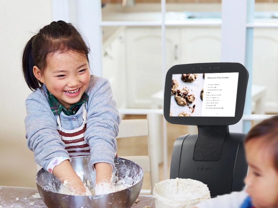 A smiling young child puts her hands into a bowl of batter. Temi stands next to her, displaying a cookie recipe.