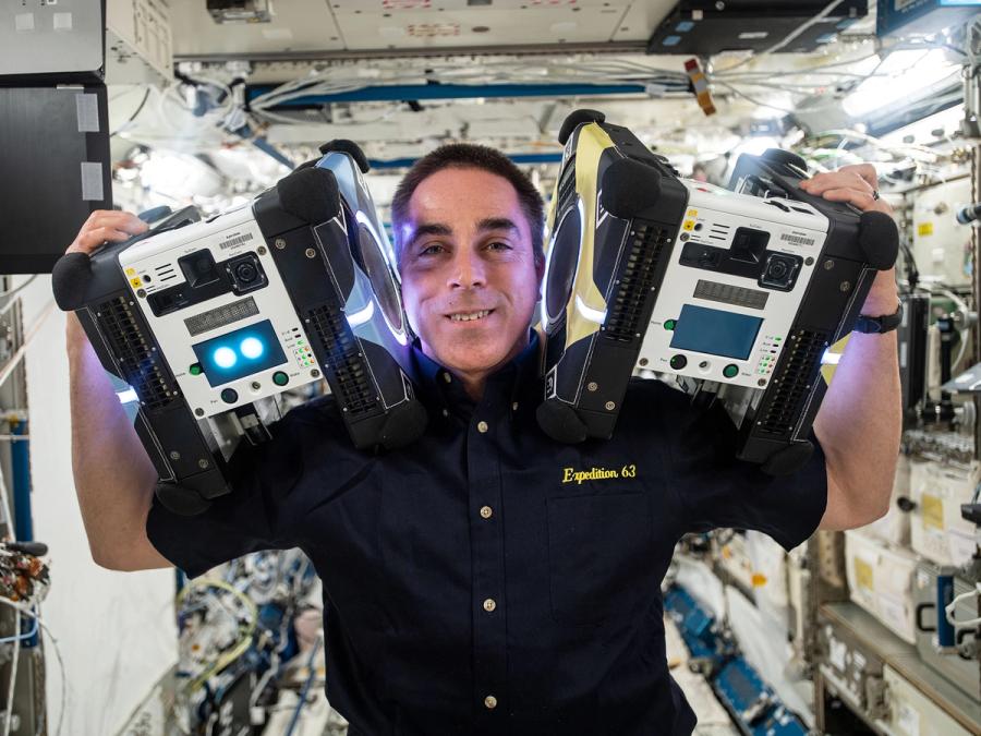 A smiling astronaut on a space station holds two square robots that are larger then his head up on each of his shoulders.