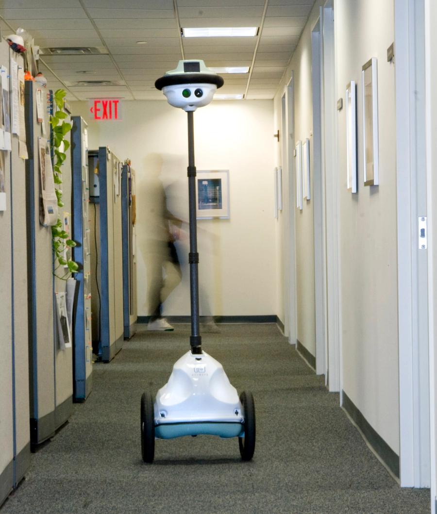 A wheeled telepresence robot goes down a corridor of offices and cubicles.