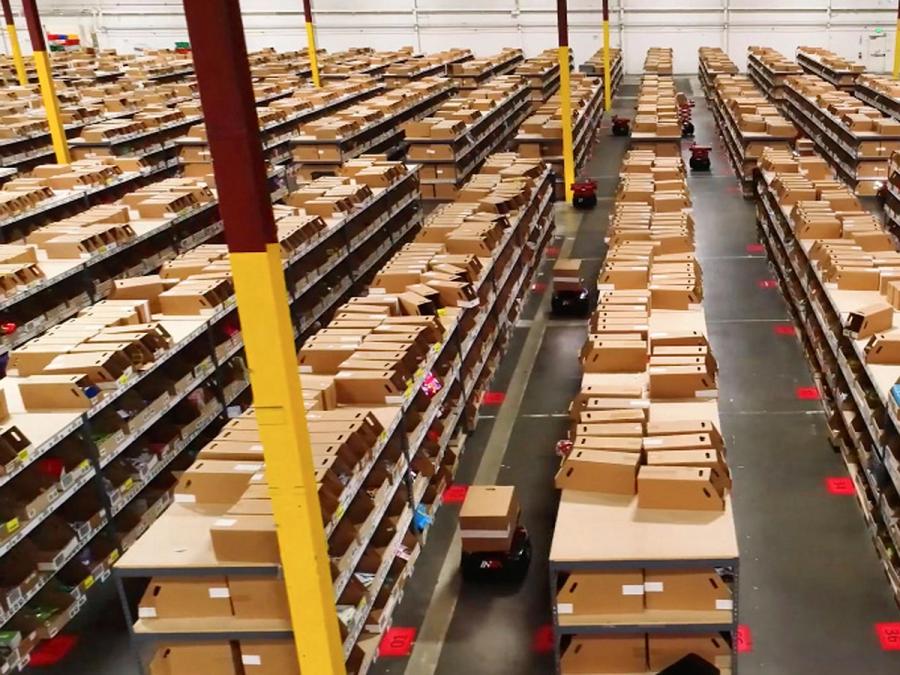 Overhead view of a warehouse with many rows of shelves with packages is seen with squat Picker Robots carrying boxes through the aisles.