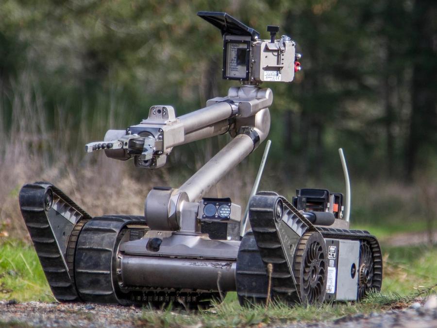 A base with large tracked wheels supports a multi-jointed tall arm which has a gripper hand at one joint, and a camera system at the end.