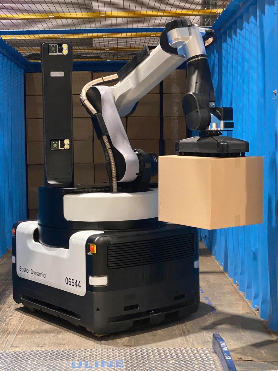 The Stretch robot sits in a blue shipping container, holding a cardboard box by the vacuum gripper at the end of its arm.