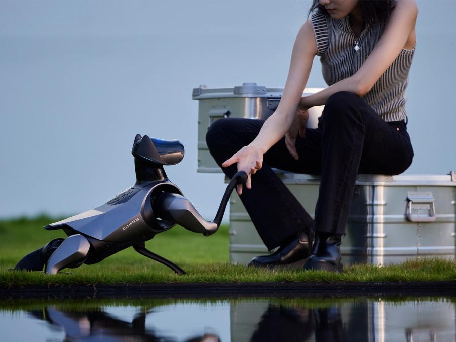 A black and silver Xiaomi CyberDog 2 robotic dog puts it's paw into the hand of a woman sitting on a silver trunk.