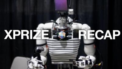 A humanoid robot with a screen for face, white plastic arms and hands, black and white shirt, stand against a black background.