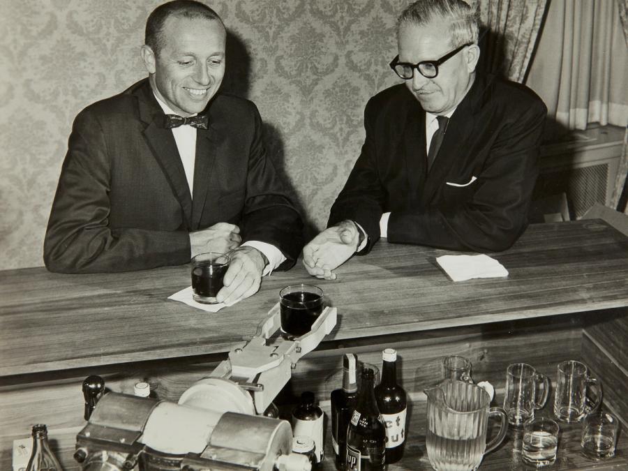 Black and white photo of two men at a bar being served drinks by a robotic arm 