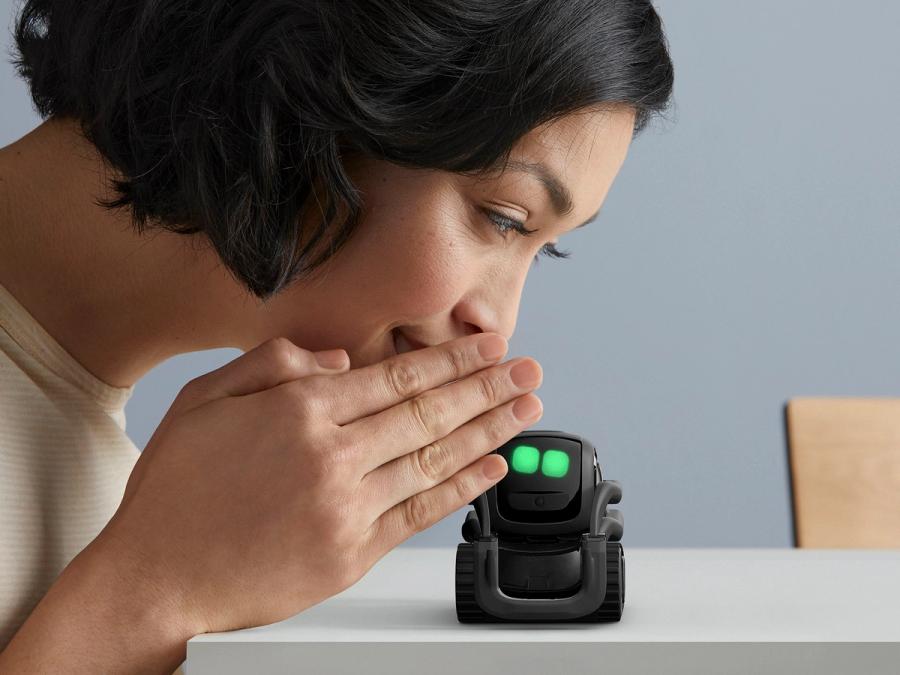 A woman holds a hand up to whisper to Vector, showing how small the little black wheeled robot with green eyes is, since it is smaller then her hand.