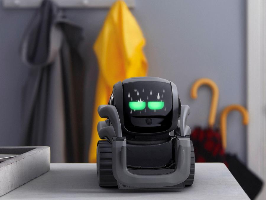 The Vector robot is a simple, compact, black wheeled robot smaller then the palm of a hand with two glowing green eyes sits on surface in a home.