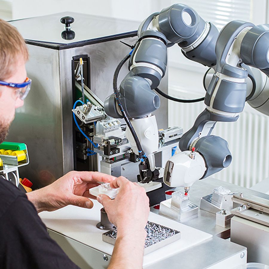 A white base and torso with two industrial arms works with a person wearing safety glasses, and holding plastic piece in his hands, in a factory setting.