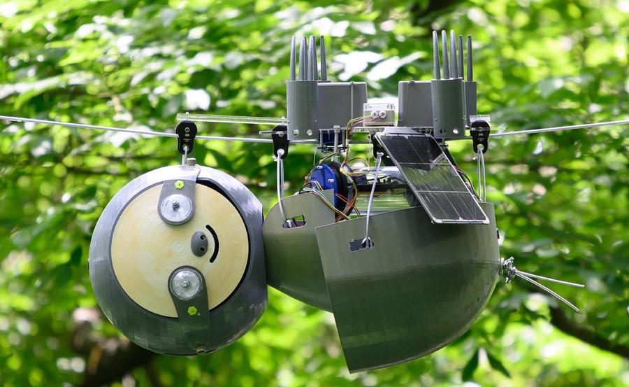 The SlothBot robotic sloth hangs upside down from a wire amid green leafy trees outside. It has a solar panel and other electronics.