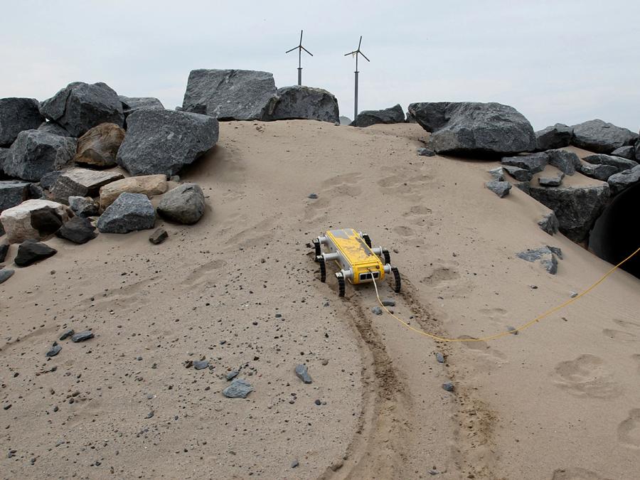 A low to the ground six legged robot walks on beachy sand.