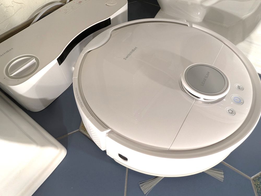 A round white robot vacuum docks to a rectangular white dock on a bathroom floor.