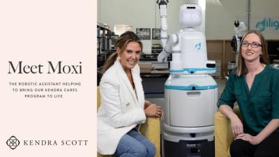 Two women smile at the camera sitting next to Moxi robot.