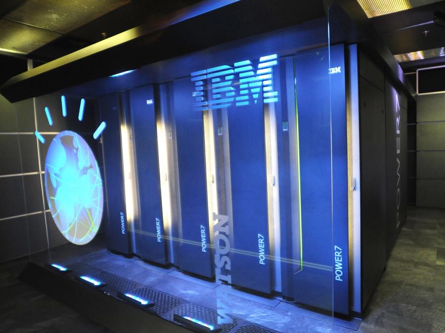 A room is filled with a large floor to ceiling boxy shape consisting of connected server racks labelled Power7. A glass wall at the front says IBM Watson and shows a logo with a blue circle and lines on top.