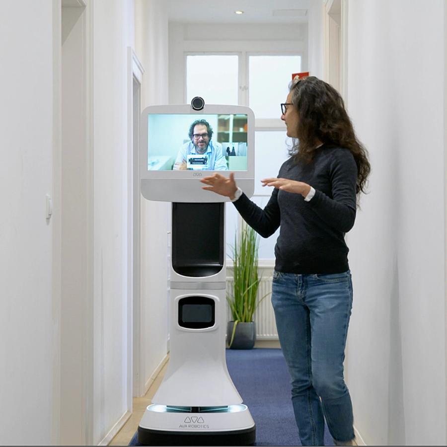 A woman walks down a hallway talking to a man seen on the screen of a telepresence robot.