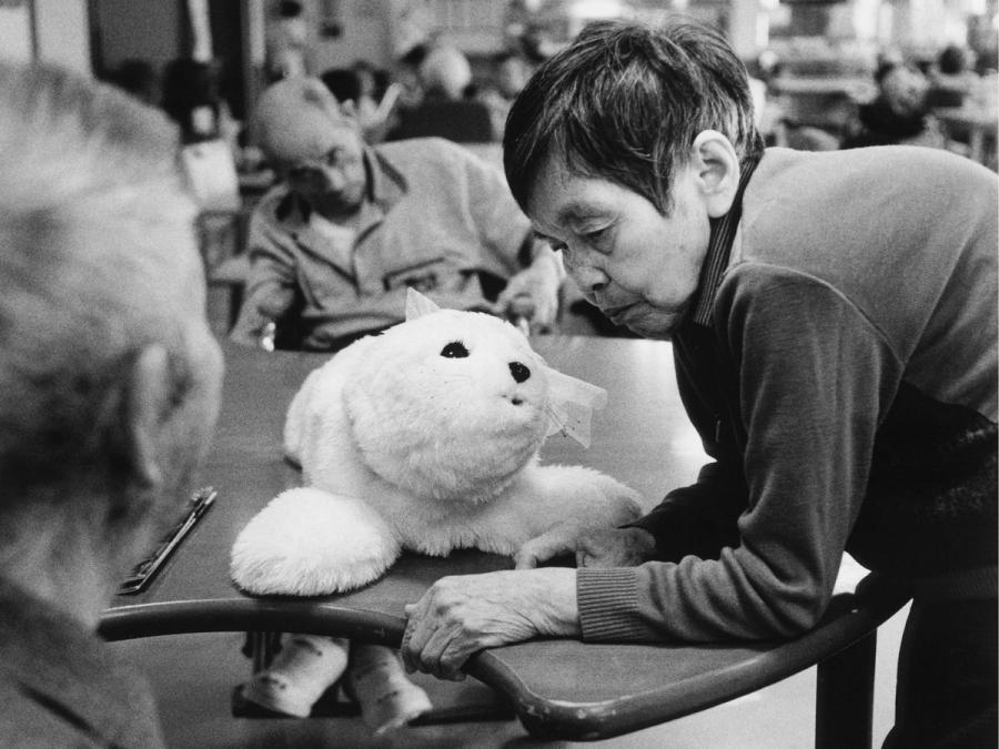 An elderly woman presses her face towards a white robotic seal on the table at a nursing home.