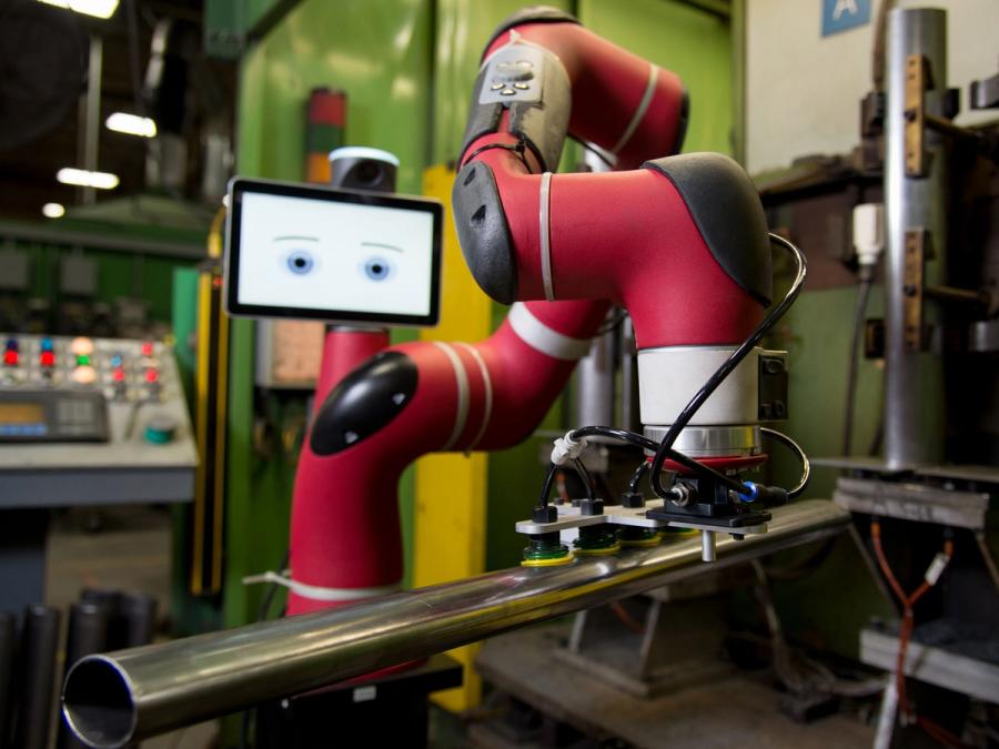 An industrial one armed robot with a tablet display for a head picks up a metal pipe.