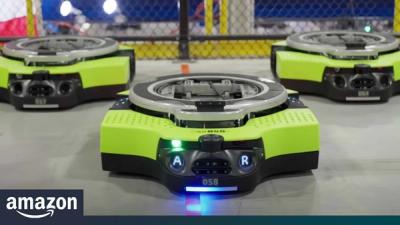 A green, gray, and black squat robot designed to carry products on its back stands on a concrete floor next to two other identical robots on an Amazon warehouse.