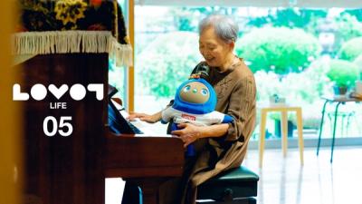 An older woman smiles while sitting at a piano with LOVOT on her lap.