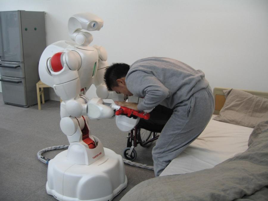 A large white humanoid on a mobile base helps an adult male who is bent over trying to get out of bed.