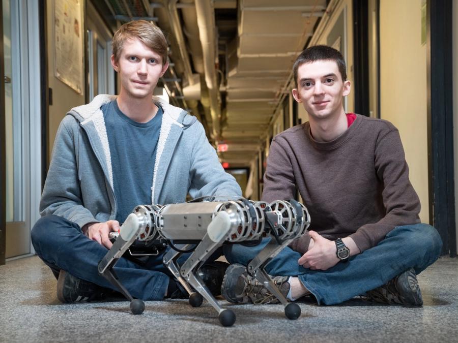 Two men sit behind a small four legged robot. The scale shows the robot as coming up to their knees while seated.