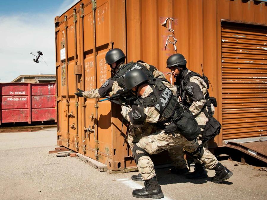 Three SWAT members in gear and camouflage gather behind a wall as one tosses a Throwbot.